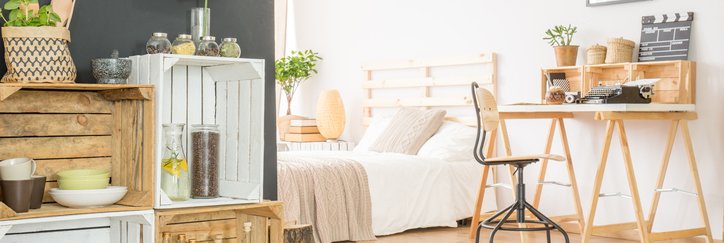 A sunny studio apartment with a desk, low bed, and wooden crates for shelves,