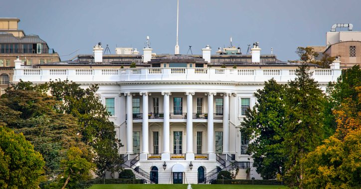 The White House surrounded by trees and a lawn with the American flag flying at the top of the building.