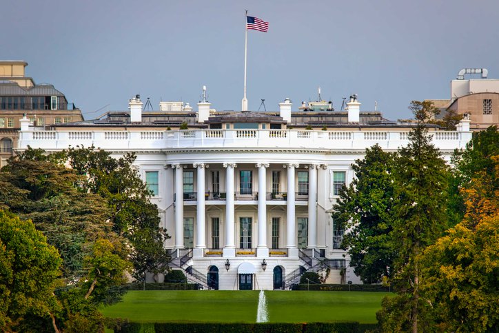 The White House surrounded by trees and a lawn with the American flag flying at the top of the building.