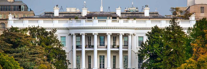 The White House surrounded by trees and a lawn with the American flag flying at the top of the building.