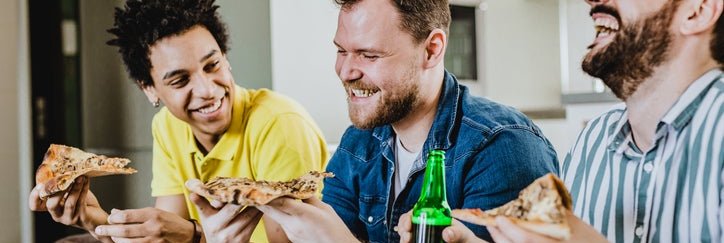 Three male friends laughing while eating pizza at home on the couch.