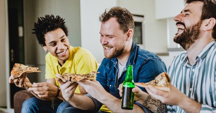 Three male friends laughing while eating pizza at home on the couch.