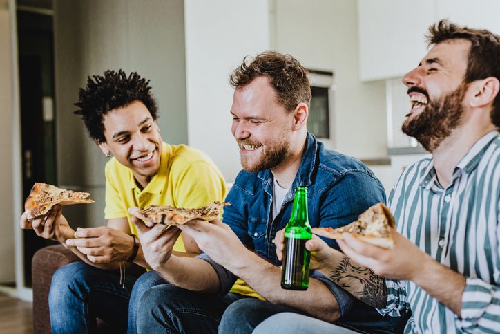 Three male friends laughing while eating pizza at home on the couch.