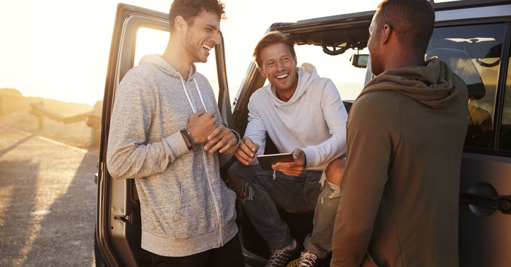 Three smiling men standing around their car and planning their road trip route on a phone.