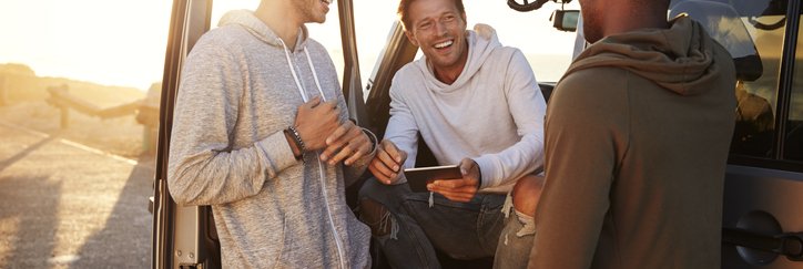 Three smiling men standing around their car and planning their road trip route on a phone.