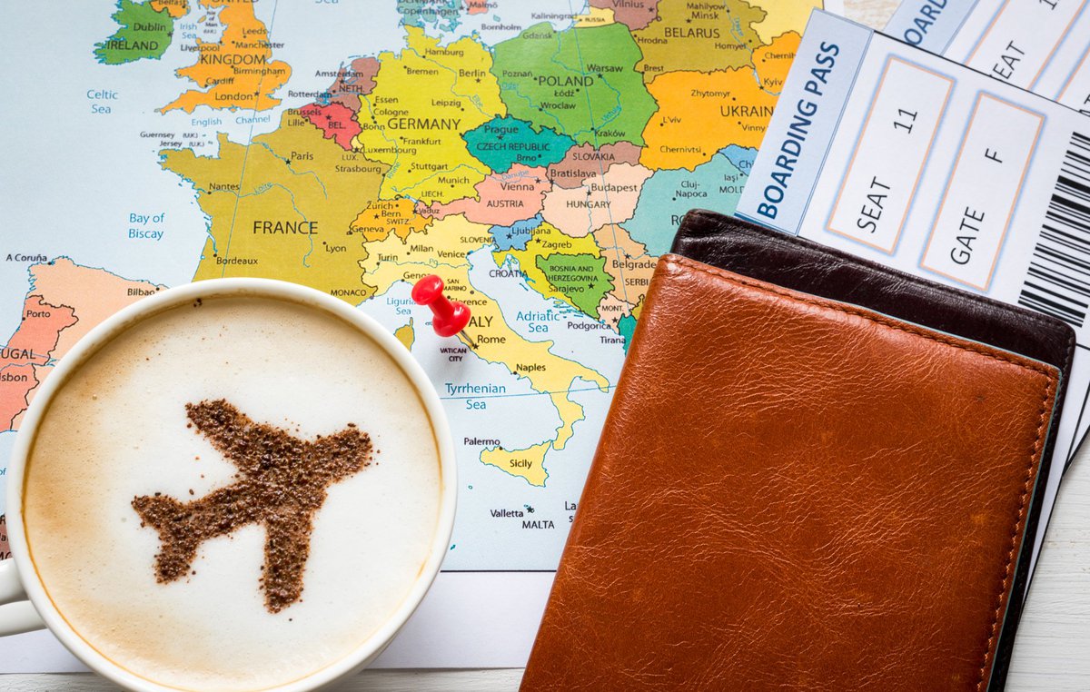 Cup of coffee with plane image in it sitting on top of map and next to a boarding pass.