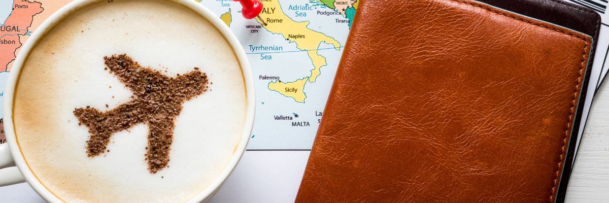 Cup of coffee with plane image in it sitting on top of map and next to a boarding pass.