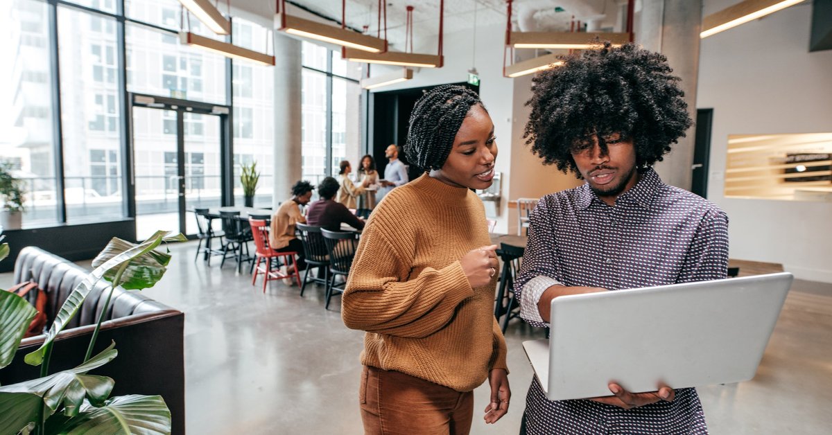 Two coworkers standing in an open office and looking at a laptop one of them is holding.