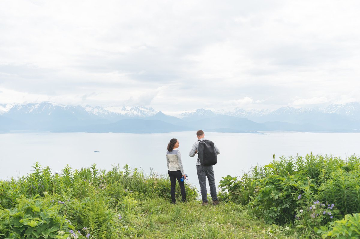 Two hikers at a grassy lookout point with water and mountains in the distance.