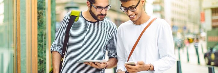 Two men standing on a sunny street, one holding a tablet, the other holding a phone and credit card.