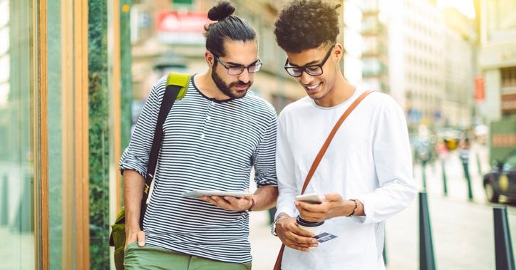 Two men standing on a sunny street, one holding a tablet, the other holding a phone and credit card.