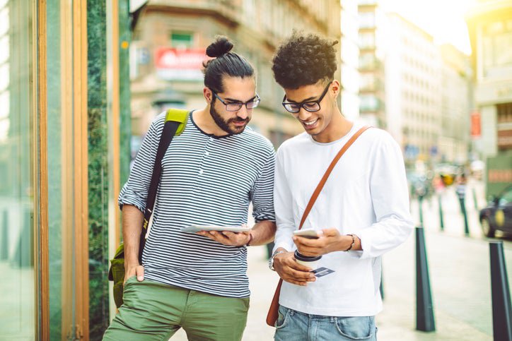 Two men standing on a sunny street, one holding a tablet, the other holding a phone and credit card.
