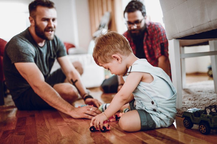 Two fathers playing with toy cars on the floor with their toddler son.