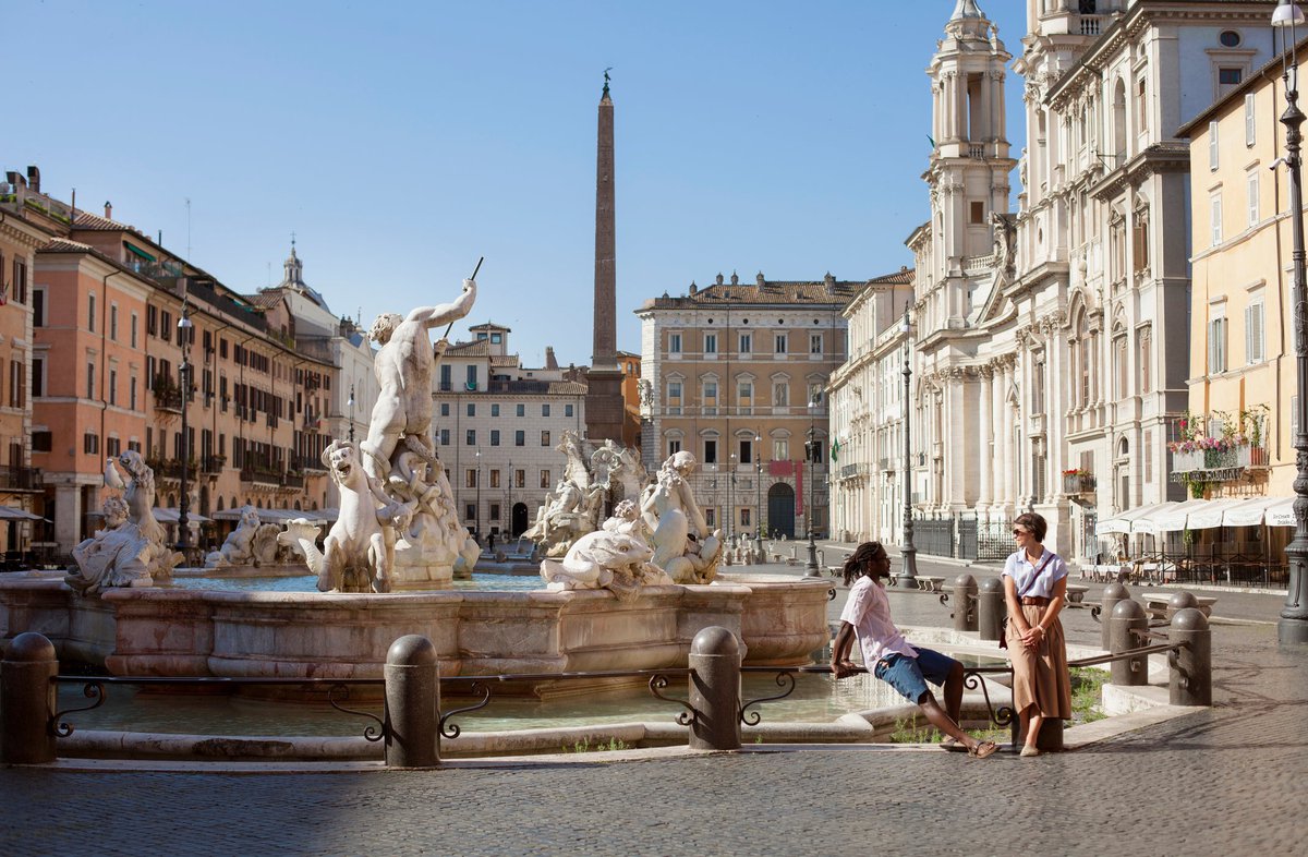 Two vacationers sitting next to a beautiful fountain in an open square in Rome, Italy.