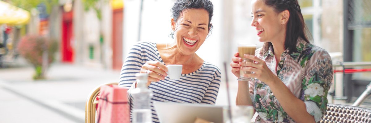 Two smiling people sipping coffee at an outdoor cafe table next to a shopping bag.