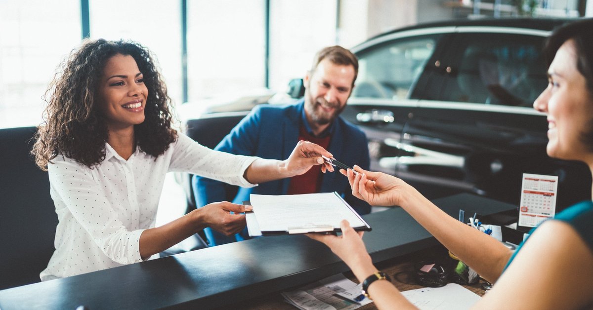 Two people sitting at a desk in a car showroom and handing signed forms back to the salesperson.