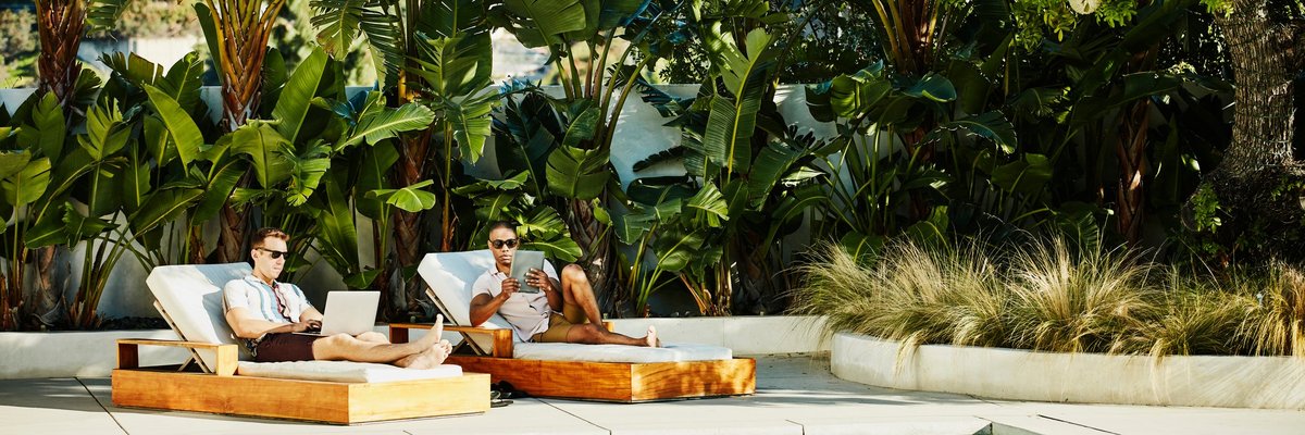 Two people lounging next to a luxury pool surrounded by palm trees while typing on a laptop and tablet.