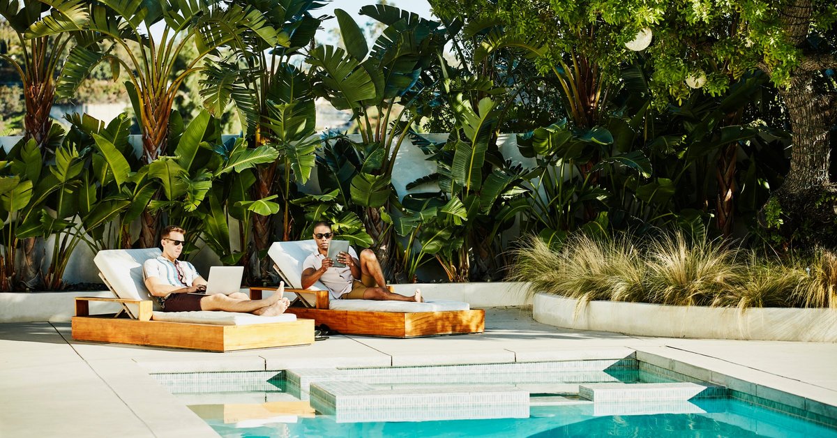 Two people lounging next to a luxury pool surrounded by palm trees while typing on a laptop and tablet.