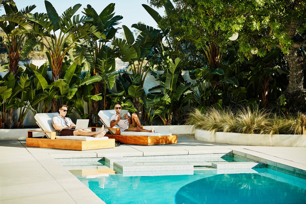 Two people lounging next to a luxury pool surrounded by palm trees while typing on a laptop and tablet.