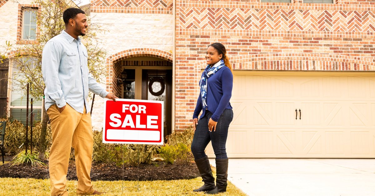 Two people standing next to a For Sale sign in front of a house.