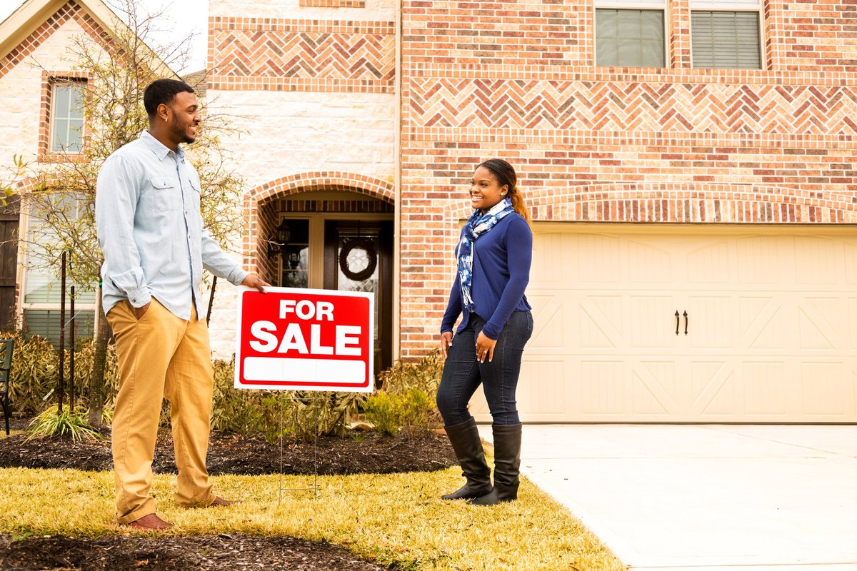 Two people standing next to a For Sale sign in front of a house.