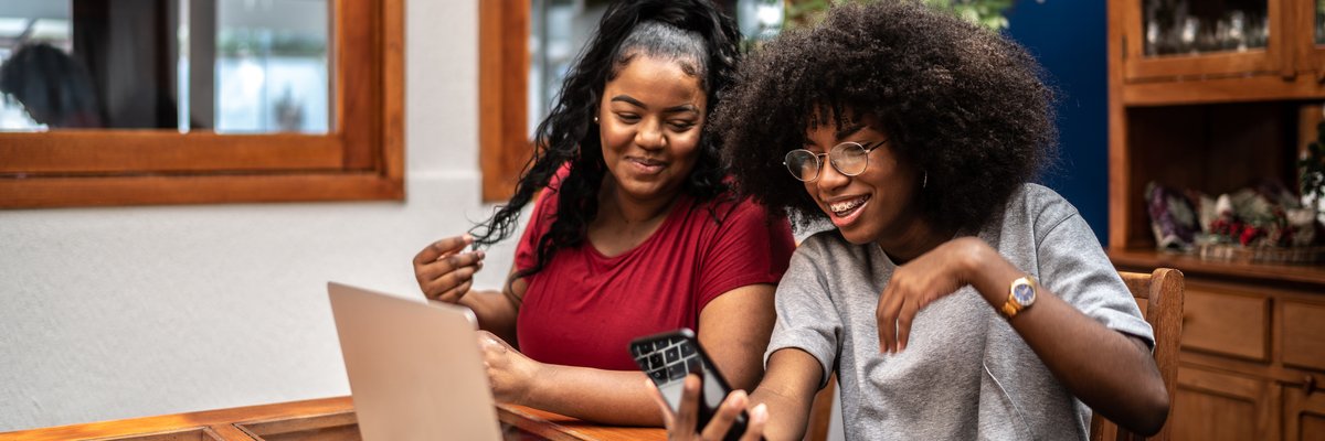 Two smiling people sitting in their kitchen and looking at a laptop while one holds a phone.