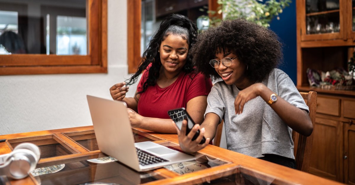 Two smiling people sitting in their kitchen and looking at a laptop while one holds a phone.