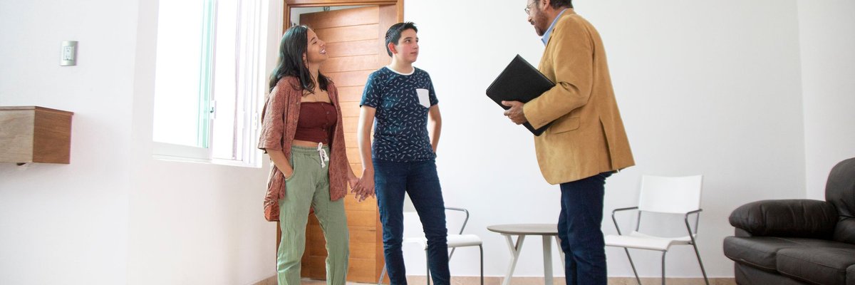 Two people holding hands while standing in the living room of a new house and talking with a realtor.