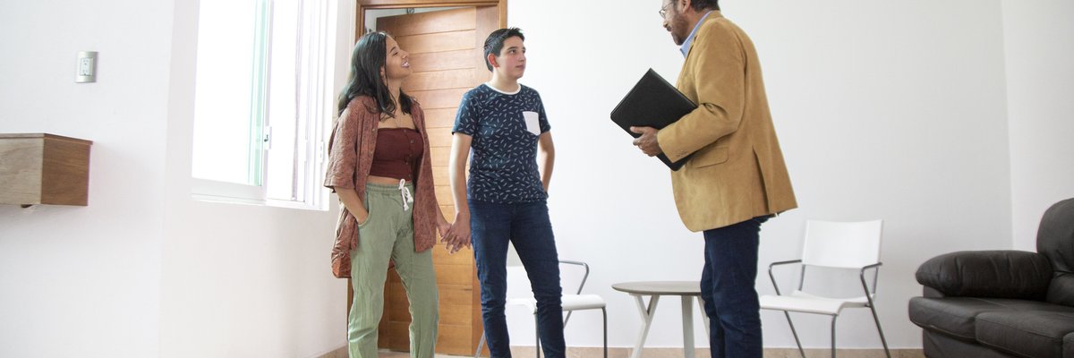 Two people holding hands while standing in the living room of a new house and talking with a realtor.