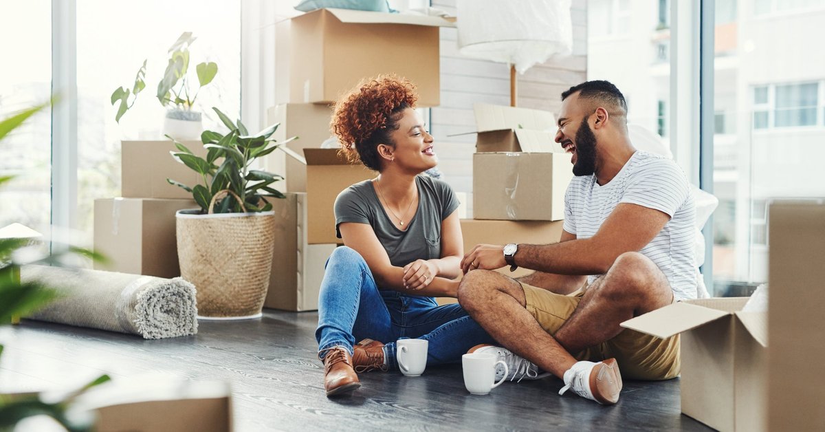 Two smiling people sitting on the floor between moving boxes.