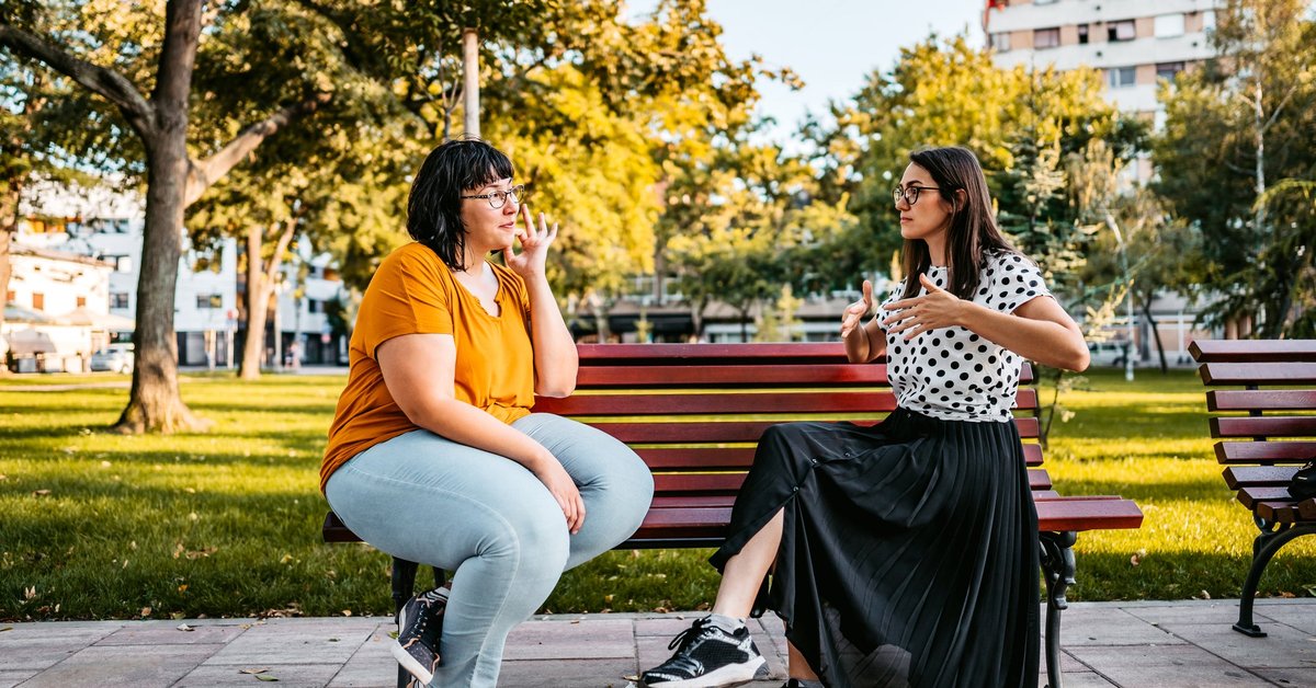 Two people sitting on a park bench and speaking in sign language.