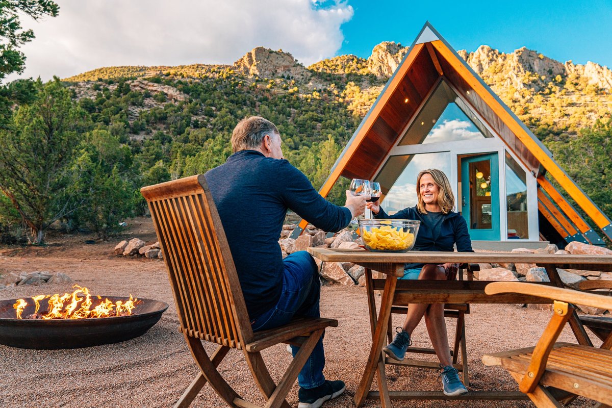Two people sitting at a dining table next to a fire pit outside their A-frame tiny home in with trees and mountains in the background.