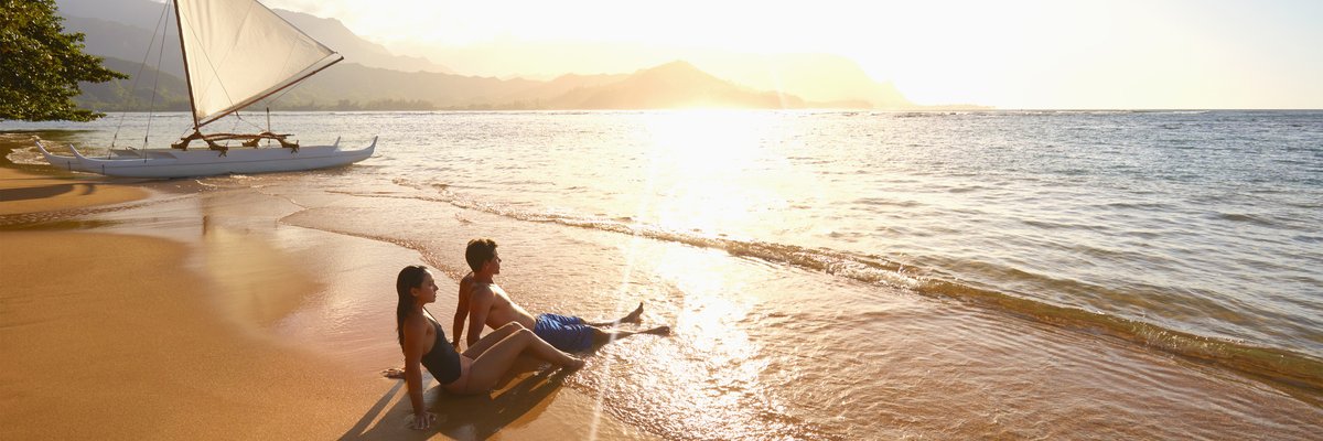 Two people sitting in the waves on a tropical beach at sunset with a sailboat behind them.