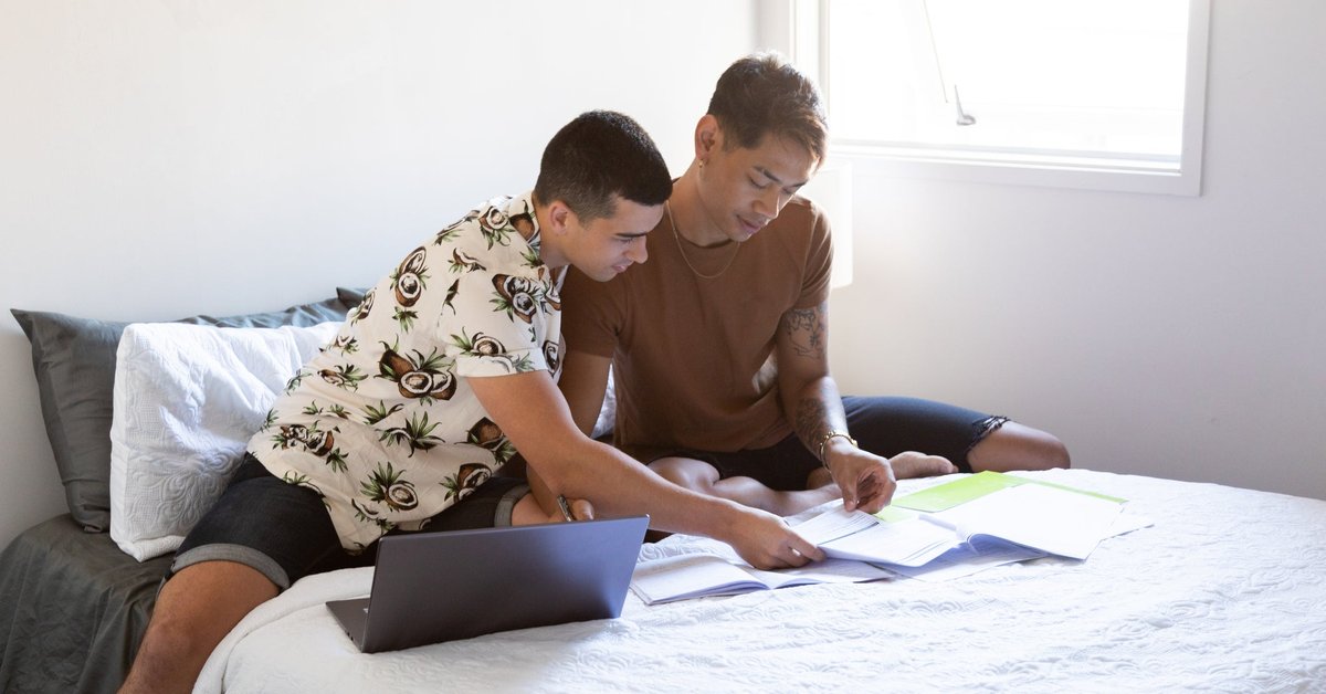 Two people sitting on a bed and looking at paperwork next to an open laptop.