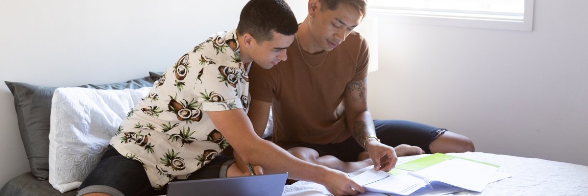 Two people sitting on a bed and looking at paperwork next to an open laptop.