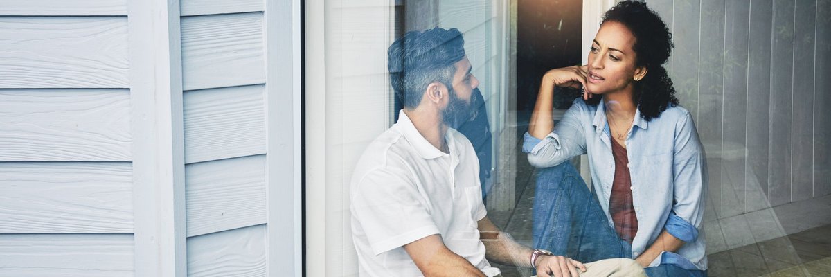 Two people sitting on the floor and talking next to a large living room window.