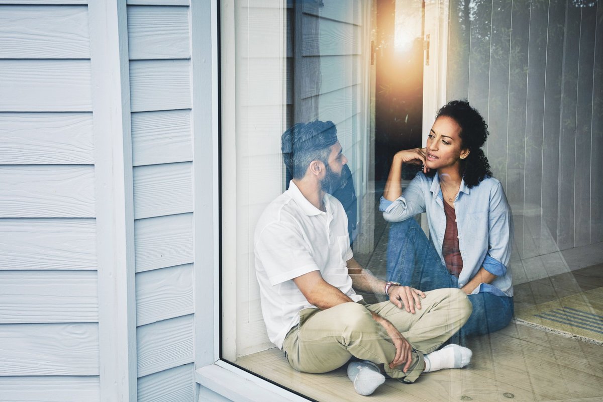 Two people sitting on the floor and talking next to a large living room window.