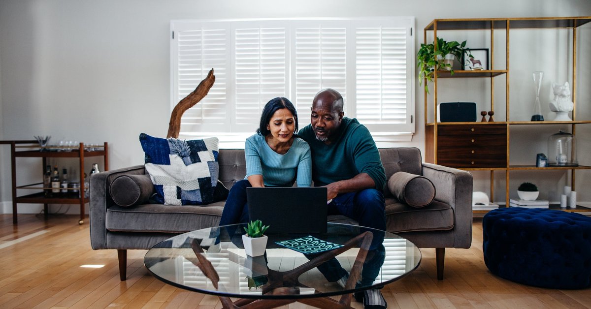 Two people sitting on a couch in their living room while typing on a laptop on the coffee table.