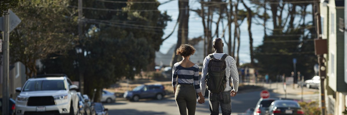 Two people holding hands while walking down the middle of a street surrounded by trees and parked cars.