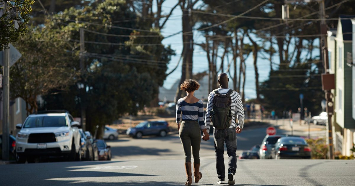 Two people holding hands while walking down the middle of a street surrounded by trees and parked cars.