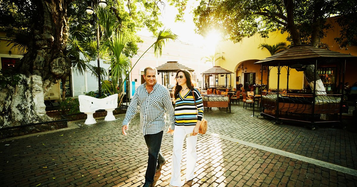 Two smiling people holding hands and walking through a sunny courtyard lined with trees.