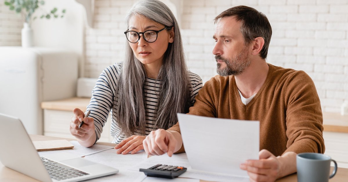 Two people sitting at their kitchen table with an open laptop, paperwork, and a calculator.