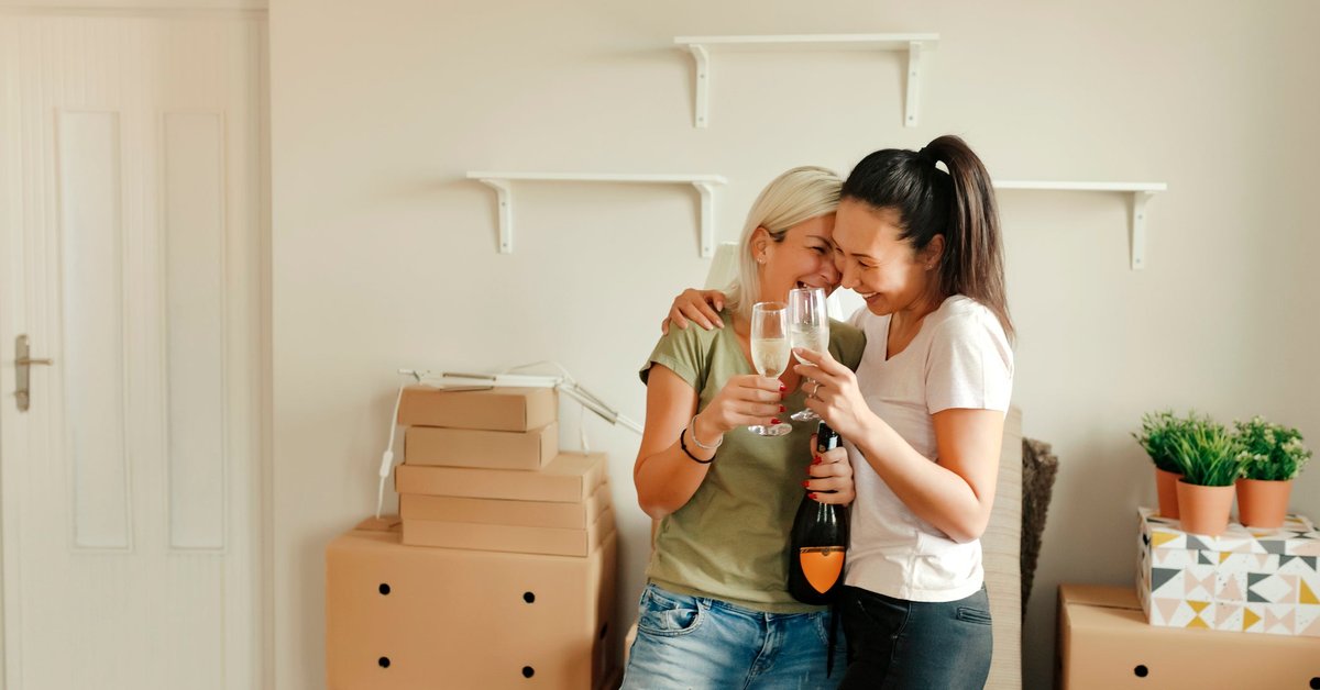 Two smiling people standing in a living room surrounded by moving boxes and drinking sparkling wine.