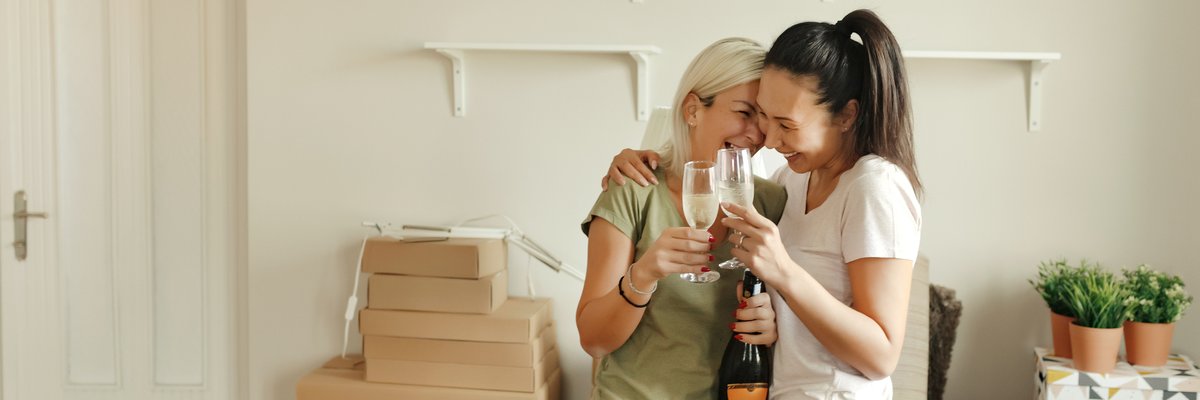 Two smiling people standing in a living room surrounded by moving boxes and drinking sparkling wine.