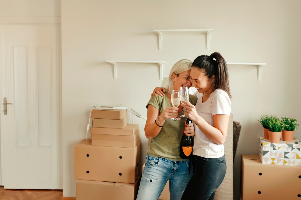 Two smiling people standing in a living room surrounded by moving boxes and drinking sparkling wine.