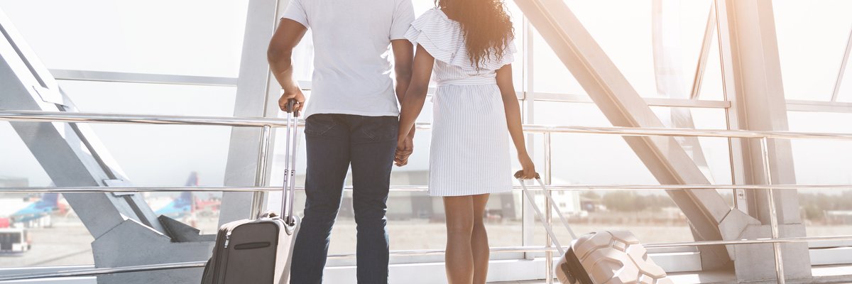 Two people standing next to their suitcases at an airport window and holding hands.