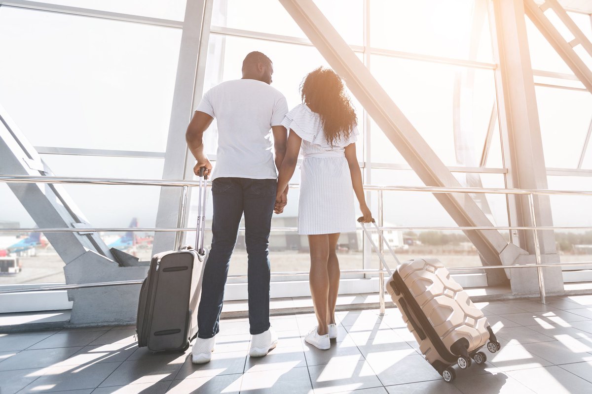 Two people standing next to their suitcases at an airport window and holding hands.