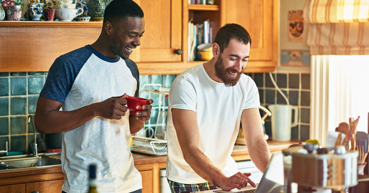 Two roommates laughing while watching something on a computer on the kitchen counter.
