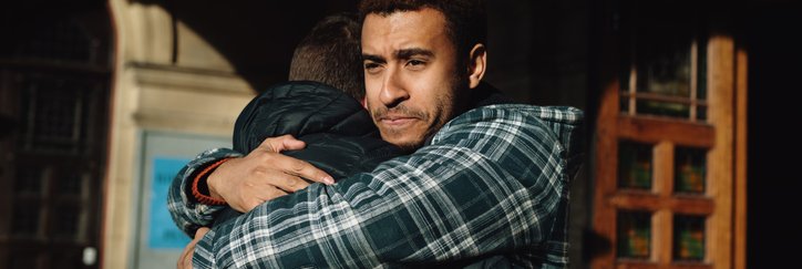 A man looking sad while hugging another man outside a doorway.