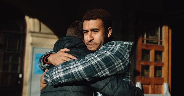 A man looking sad while hugging another man outside a doorway.